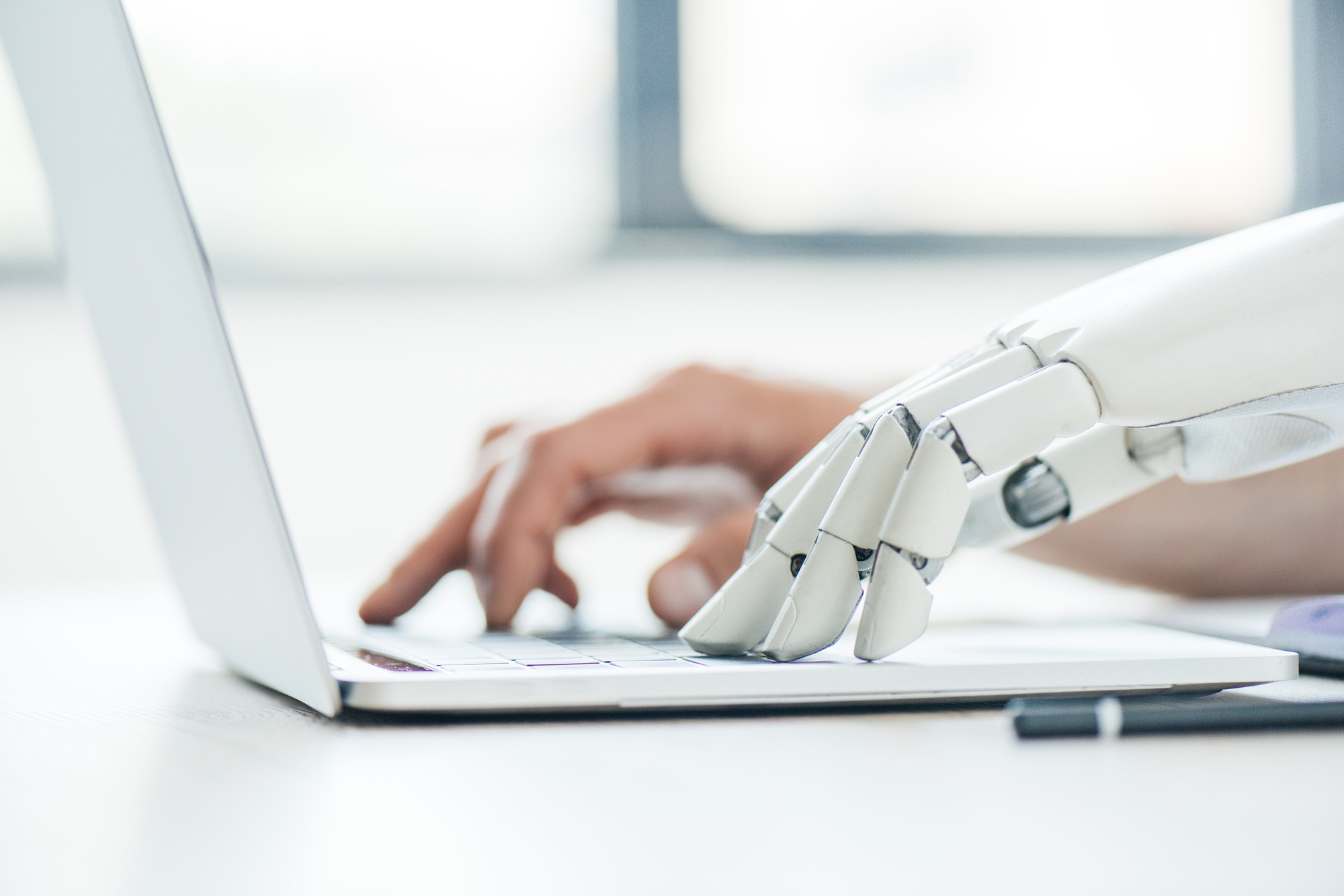 selective focus of human and robot hands typing on laptop at workplace